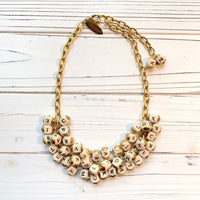 May include: A gold chain necklace with white letter beads. The beads are arranged in a cluster and spell out the letters of the alphabet. The necklace is on a white wooden surface. Lenora Dame necklace.