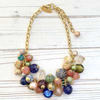 May include: Gold chain necklace featuring a cluster of colorful beads. Beads include various textures and patterns: clear, frosted, glittery, striped, and solid colors such as peach, teal, green, blue, brown, and cream. The necklace has a gold clasp. Lenora Dame necklace.