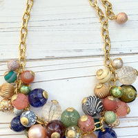 Gold chain necklace with multicolored beads. The beads are various shapes and sizes, including round, faceted, and textured. Colors include pink, peach, green, blue, brown, and clear. Some beads have gold accents. The necklace has a gold clasp with a small tag. The beads appear to be glass or resin.