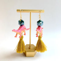 Cute dinosaur earrings for women with miniature pink dinosaurs topped with a dark green faceted bead, and a gold silk thread tassel hanging below. Dino earrings.