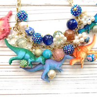 A whimsical statement necklace features a gold chain adorned with colorful beads, pearls, and miniature dinosaur figurines. The beads include shades of pink, orange, teal, blue, and purple, creating a vibrant and playful aesthetic. The necklace showcases a fun, eclectic mix of textures and materials, making it a unique accessory.
