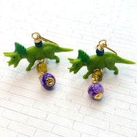 Miniature triceratops earrings with miniature green triceratops figurines dangling from gold lever back and one yellow bead and one purple bead dangling below the dinosaurs. Cute dinosaur earrings. Lenora Dame dinosaur jewelry.