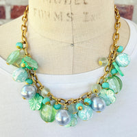 A mix of light green and light blue beads, pearls, and mother of pearl flowers hand from a gold chain necklace shown on a dress form wearing a white shirt. Green beaded necklace for women made in the USA. Lenora Dame necklace.