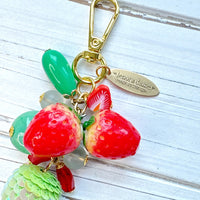 Cute strawberry bag charm with a mix of red and green beads, two red strawberry charms on a gold chain with gold trigger clasp and brass hang tag that says Lenora Dame Made in USA. Lenora Dame bag charm.