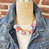 Chunky charm necklace with assorted mix of light blue seed bead balls, floral fimo beads, blue and orange mother of pearl disc bead hung on a gold necklace chain with lobster clasp and hangtag that says Lenora Dame Made in the USA. Necklace is shown on a dress form wearing a white shirt and blue denim jacket with a red brick wall background. Lenora Dame necklace.