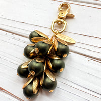 Gold bag charm with a trigger clasp at the top holding a cluster of matte hunter green pearls encased in gold leaf bead caps. A gold oval tag imprinted with Lenora Dame Handmade in USA hangs at the top by the trigger clasp. Lenora Dame bag charms. Green beaded purse charm.