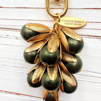 Gold bag charm with a trigger clasp at the top holding a cluster of matte hunter green pearls encased in gold leaf bead caps. A gold oval tag imprinted with Lenora Dame Handmade in USA hangs at the top by the trigger clasp. Lenora Dame bag charms. Green beaded purse charm.