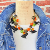 A gold chain necklace with black cat charms, orange, green, and white pumpkin charms, and black and white beads. The necklace is on a mannequin with a brick background.