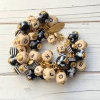 Chunky charm bracelet filled with wooden letter beads, striped square beads and round black and white beads hung on a gold cable chain with lobster clasp and Lenora Dame hang tag.