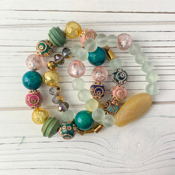 Stretch bracelet set of three bracelets with green, pink, gold, and blue beads. The bracelets are made of glass and metal beads and are arranged in a circle on a white wooden surface. Stacking bracelets for women. Lenora Dame bracelets.