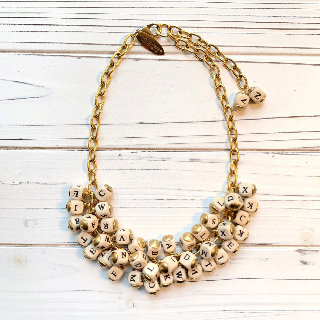 May include: A gold chain necklace with white letter beads. The beads are arranged in a cluster and spell out the letters of the alphabet. The necklace is on a white wooden surface. Lenora Dame necklace.