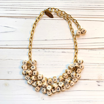 May include: A gold chain necklace with white letter beads. The beads are arranged in a cluster and spell out the letters of the alphabet. The necklace is on a white wooden surface. Lenora Dame necklace.