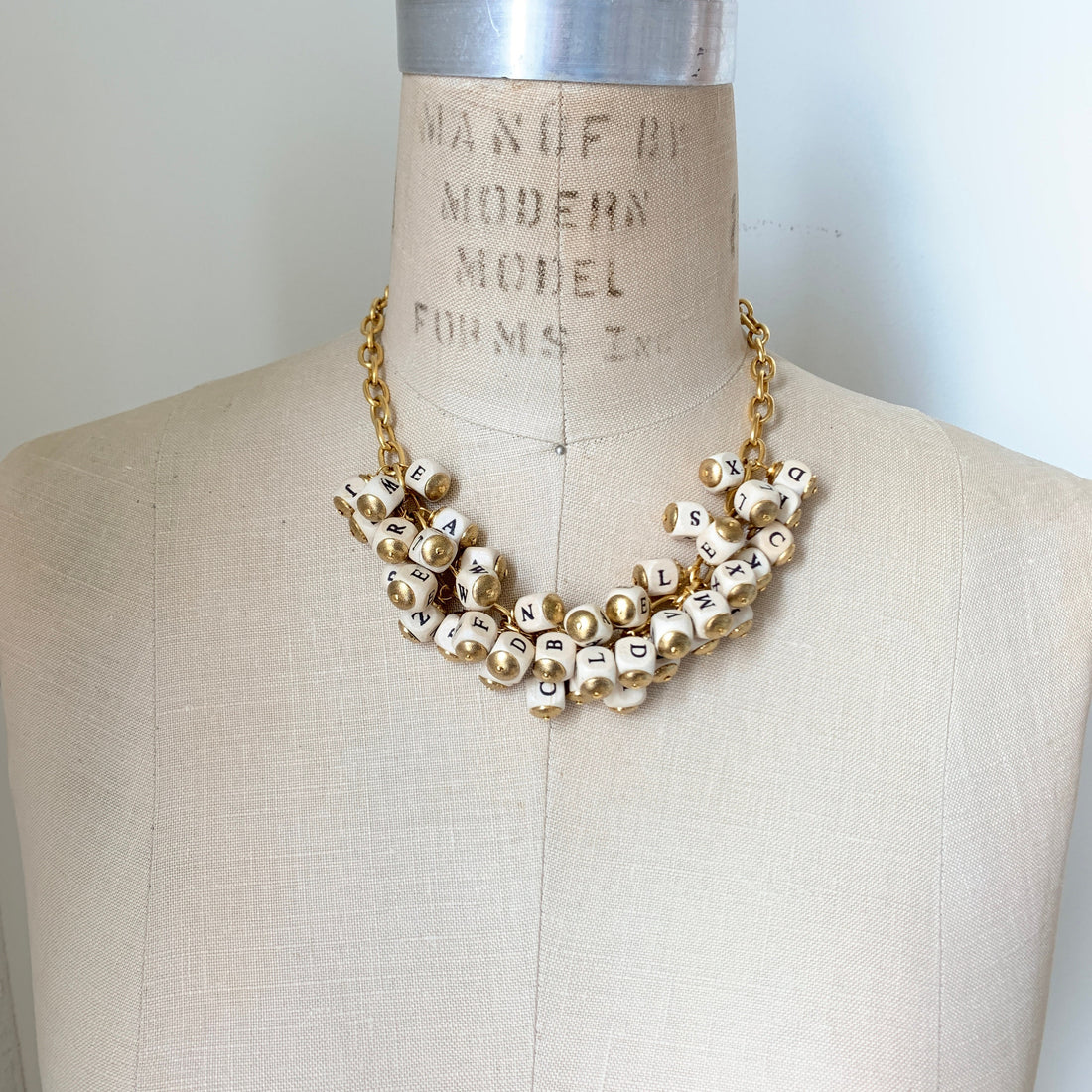 May include: A gold chain necklace with white beads that have black letters on them. The letters spell out the alphabet. The necklace is displayed on a white mannequin. Lenora Dame necklace.