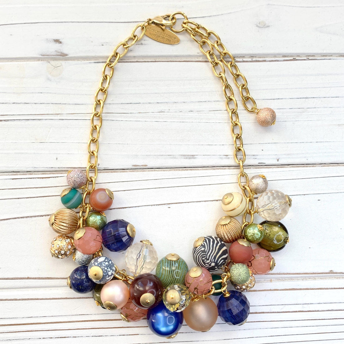 May include: Gold chain necklace featuring a cluster of colorful beads. Beads include various textures and patterns:  clear, frosted, glittery, striped, and solid colors such as peach, teal, green, blue, brown, and cream.  The necklace has a gold clasp. Lenora Dame necklace.