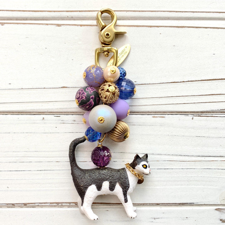 Cute bag charm for handbag with a mix of purple and gold beds on a gold chain and a black and white cat with chain collar hanging at the bottom. Lenora Dame bag charm. Beaded purse charm.