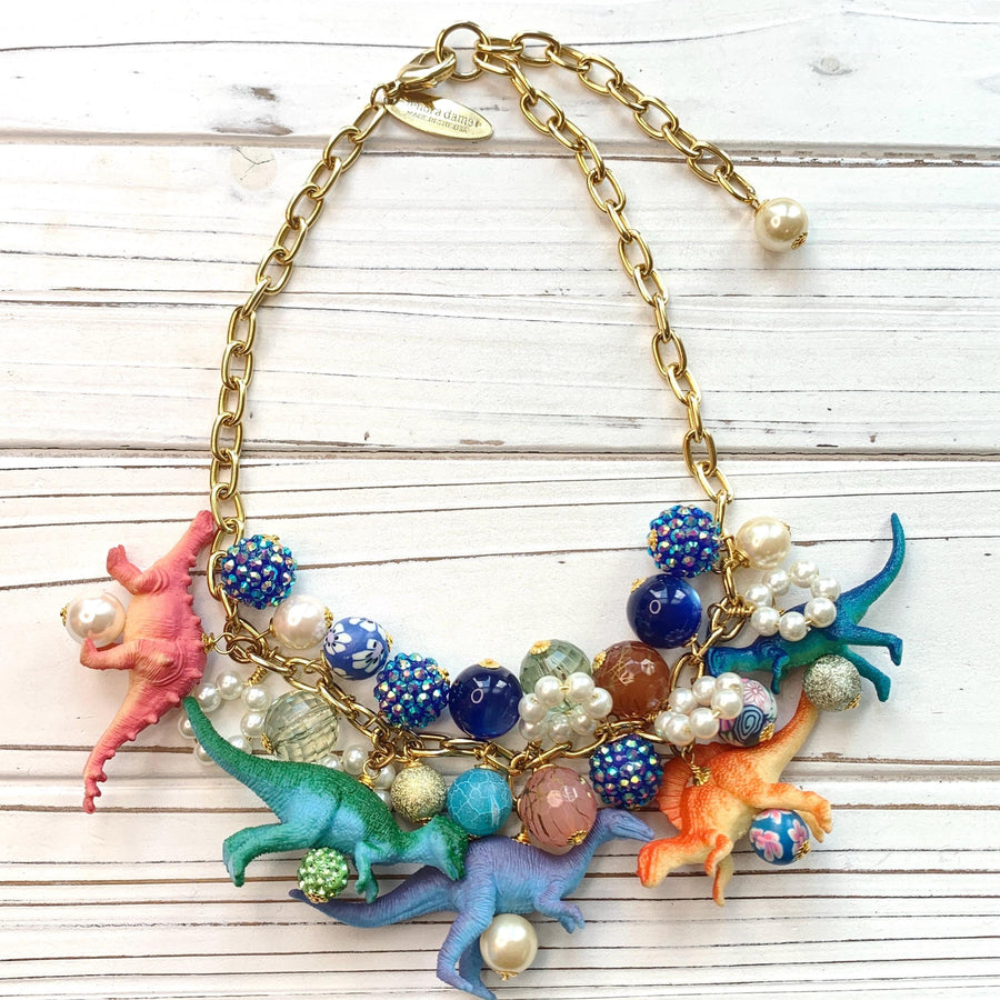A whimsical statement necklace features a gold chain adorned with colorful beads, pearls, and miniature dinosaur figurines. The beads include shades of pink, orange, teal, blue, and purple, creating a vibrant and playful aesthetic.  The necklace showcases a fun, eclectic mix of textures and materials, making it a unique accessory.