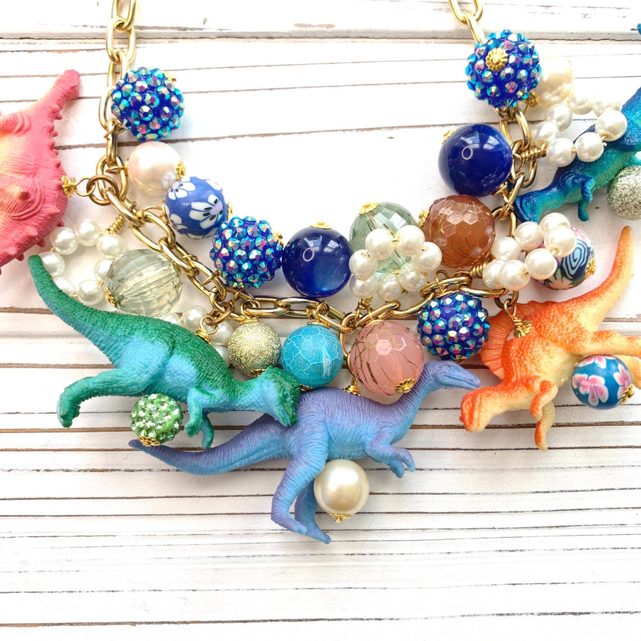 A whimsical statement necklace features a gold chain adorned with colorful beads, pearls, and miniature dinosaur figurines. The beads include shades of pink, orange, teal, blue, and purple, creating a vibrant and playful aesthetic.  The necklace showcases a fun, eclectic mix of textures and materials, making it a unique accessory.