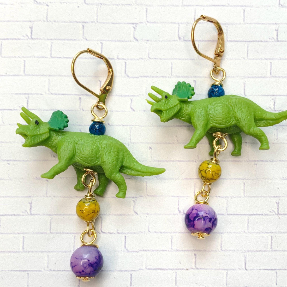 Miniature triceratops earrings with miniature green triceratops figurines dangling from gold lever back and one yellow bead and one purple bead dangling below the dinosaurs. Cute dinosaur earrings. Lenora Dame dinosaur jewelry.