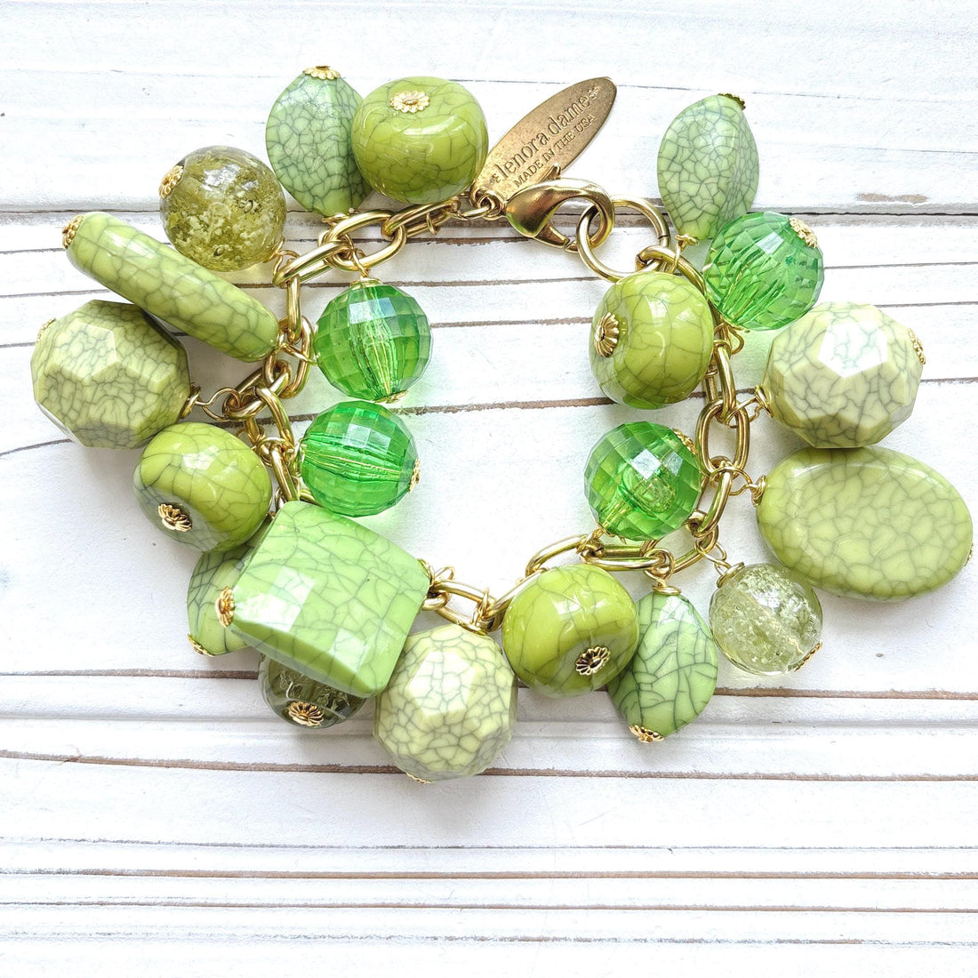 Gold chain charm bracelet filled with assorted green beads, some beads have a crackle look to them. Bracelet has a hangtag that says Lenora Dame, Handmade in the USA.