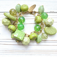 Gold chain charm bracelet filled with assorted green beads, some beads have a crackle look to them. Bracelet has a hangtag that says Lenora Dame, Handmade in the USA.