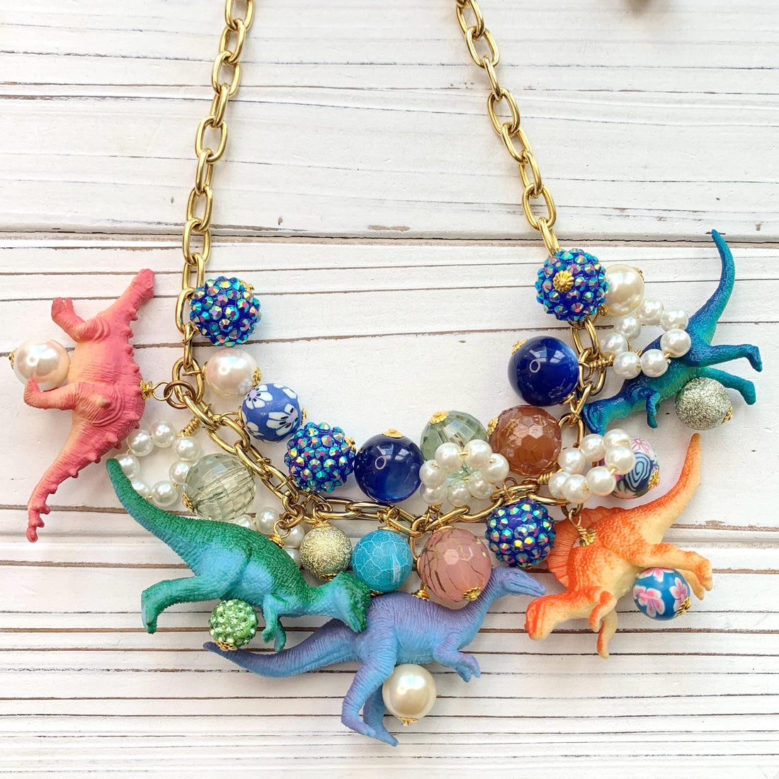 A whimsical statement necklace features a gold chain adorned with colorful beads, pearls, and miniature dinosaur figurines. The beads include shades of pink, orange, teal, blue, and purple, creating a vibrant and playful aesthetic.  The necklace showcases a fun, eclectic mix of textures and materials, making it a unique accessory.