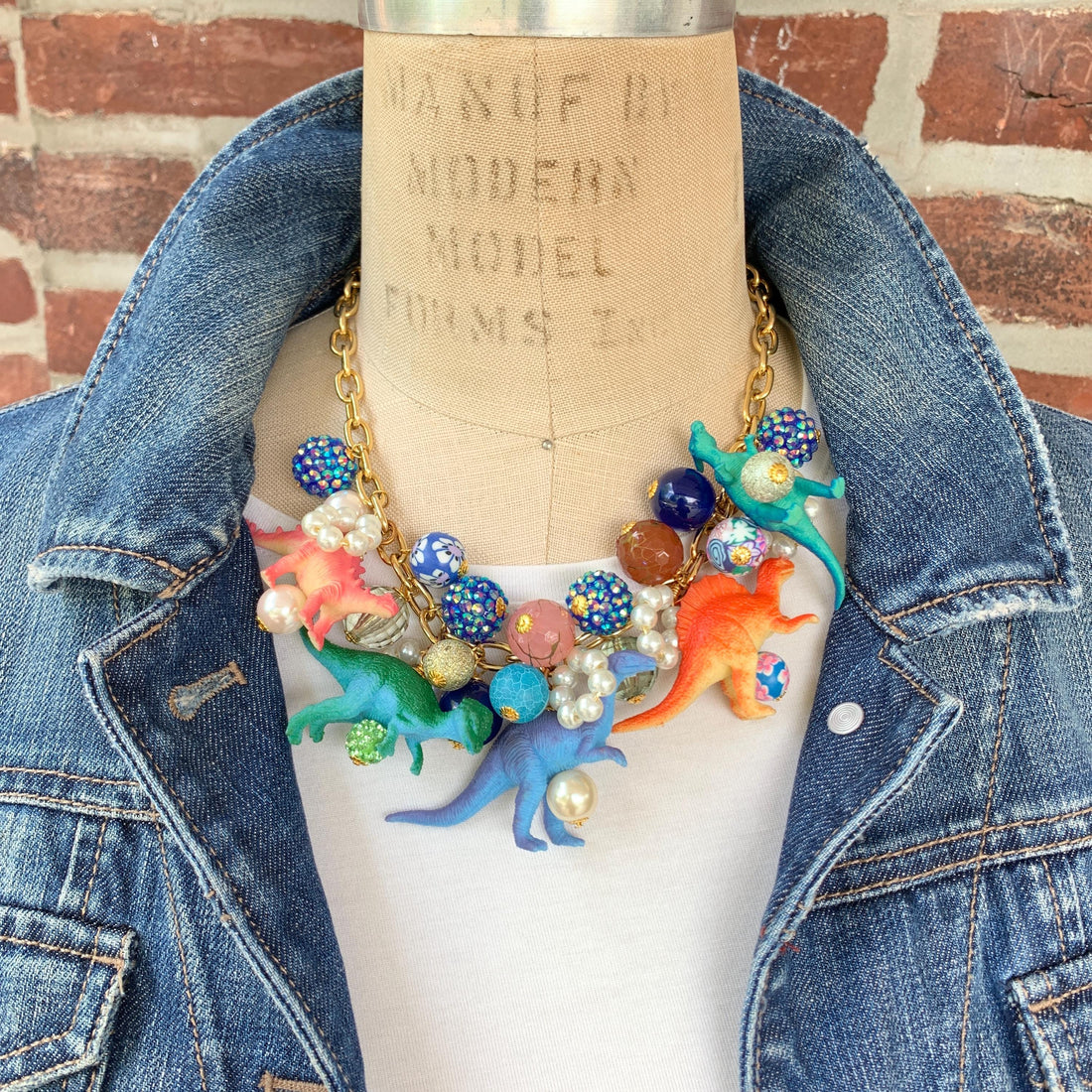 A whimsical statement necklace features a gold chain adorned with colorful beads, pearls, and miniature dinosaur figurines. The beads include shades of pink, orange, teal, blue, and purple, creating a vibrant and playful aesthetic.  The necklace showcases a fun, eclectic mix of textures and materials, making it a unique accessory.