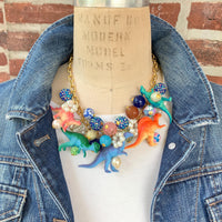 A whimsical statement necklace features a gold chain adorned with colorful beads, pearls, and miniature dinosaur figurines. The beads include shades of pink, orange, teal, blue, and purple, creating a vibrant and playful aesthetic.  The necklace showcases a fun, eclectic mix of textures and materials, making it a unique accessory.