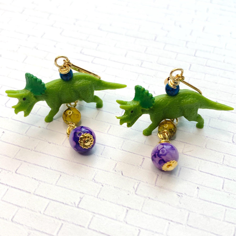 Miniature triceratops earrings with miniature green triceratops figurines dangling from gold lever back and one yellow bead and one purple bead dangling below the dinosaurs. Cute dinosaur earrings. Lenora Dame dinosaur jewelry.