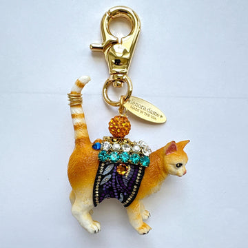 Cute cat bag charm with an orange tabby cat wearing purple and black ribbon trim embellished with green rhinestones, crystal rhinestones, and pearls. An orange rhinestone bead sits on the cat&#39;s back and is attached to the gold trigger clasp with Lenora Dame Made in USA hangtag. Lenora Dame bag charm.