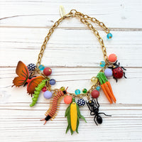 Garden bug necklace for women, with a mix of round beads, ant, butterfly, caterpillar, centipede, ladybug, corn and carrot figurines hung on a gold chain shown on a white board background. Bug jewelry for women. Lenora Dame necklace made in the USA.