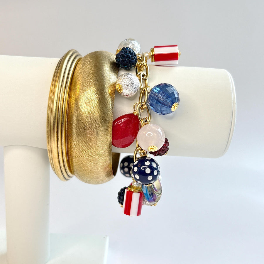 Red, white and blue charm bracelet shown on a white jewelry display arm stacked with two gold bangle bracelets. Bracelet is a mix of beads including polka dot beads, striped beads, red rhinestone beads, and blue rhinestone beads on a gold link chain. Patriotic jewelry for women. Lenora Dame jewelry.