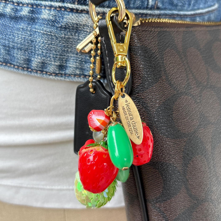 Red and green beaded bag charm with two strawberry charms, a miniature strawberry slice charm, green sequin bead, clear beads, and a long oval green bead on a gold chain with gold trigger clasp. An oval gold hang tag hangs from the bag charm and says Lenora Dame Made in USA. The bag charm is shown hung on a brown and black crossbody bag. Lenora Dame bag charm.