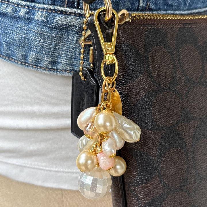 Pearl bag charm shown on a brown leather crossbody bag. A mix of cream and white faux pearls in different shapes are wire wrapped to a gold chain with gold clasp and oval hang tag that says Lenora Dame Made in USA. Lenora Dame bag charm.