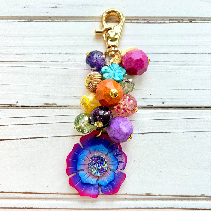 Colorful beaded bag charm filled with assorted beads in different shapes in vibrant colors. A large pink, purple and blue flower is at the bottom of the bead cluster. A gold trigger clasp is above the beads. Lenora Dame bag charms.