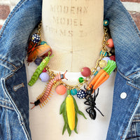 Garden bug necklace for women, with a mix of round beads, ant, butterfly, caterpillar, centipede, ladybug, corn and carrot figurines hung on a gold chain shown on a dress form wearing a white t-shirt and denim jacket. Bug jewelry for women. Lenora Dame necklace made in the USA.