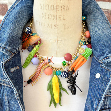 Garden bug necklace for women, with a mix of round beads, ant, butterfly, caterpillar, centipede, ladybug, corn and carrot figurines hung on a gold chain shown on a dress form wearing a white t-shirt and denim jacket. Bug jewelry for women. Lenora Dame necklace made in the USA.