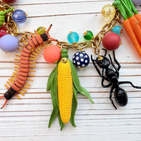 Garden bug necklace for women, with a mix of round beads, ant, butterfly, caterpillar, centipede, ladybug, corn and carrot figurines hung on a gold chain shown on a white board background. Bug jewelry for women. Lenora Dame necklace made in the USA.