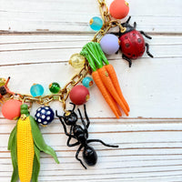 Garden bug necklace for women, with a mix of round beads, ant, butterfly, caterpillar, centipede, ladybug, corn and carrot figurines hung on a gold chain shown on a white board background. Bug jewelry for women. Lenora Dame necklace made in the USA.