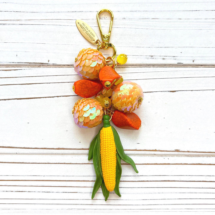 Beaded bag charm with miniature yellow corn cob with green leaves hanging from a mix of different orange beads. Gold trigger clasp at the top with an oval hang tag that says Lenora Dame Made in USA. Lenora Dame bag charm.