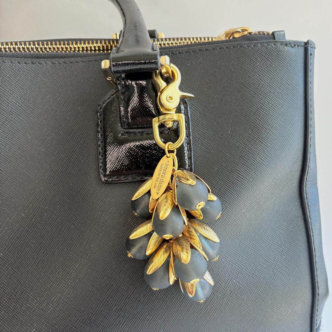 Beaded bag charm featuring matte black glass beads encased in gold plated brass floral leaf shaped bead caps. Gold trigger clasp and an oval hang tag that says Lenora Dame Made in USA. Lenora Dame bag charm shown on the strap of a black leather handbag.