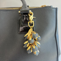 Beaded bag charm featuring matte black glass beads encased in gold plated brass floral leaf shaped bead caps. Gold trigger clasp and an oval hang tag that says Lenora Dame Made in USA. Lenora Dame bag charm shown on the strap of a black leather handbag.