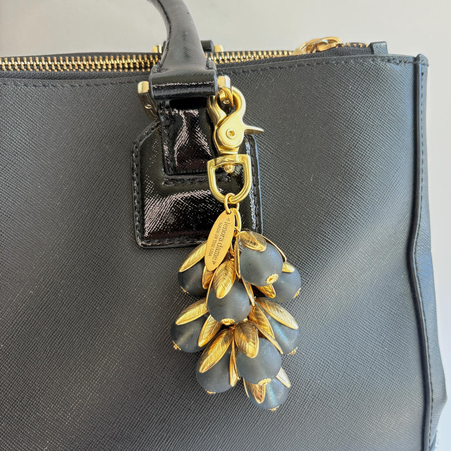 Beaded bag charm featuring matte black glass beads encased in gold plated brass floral leaf shaped bead caps. Gold trigger clasp and an oval hang tag that says Lenora Dame Made in USA. Lenora Dame bag charm shown on the strap of a black leather handbag.