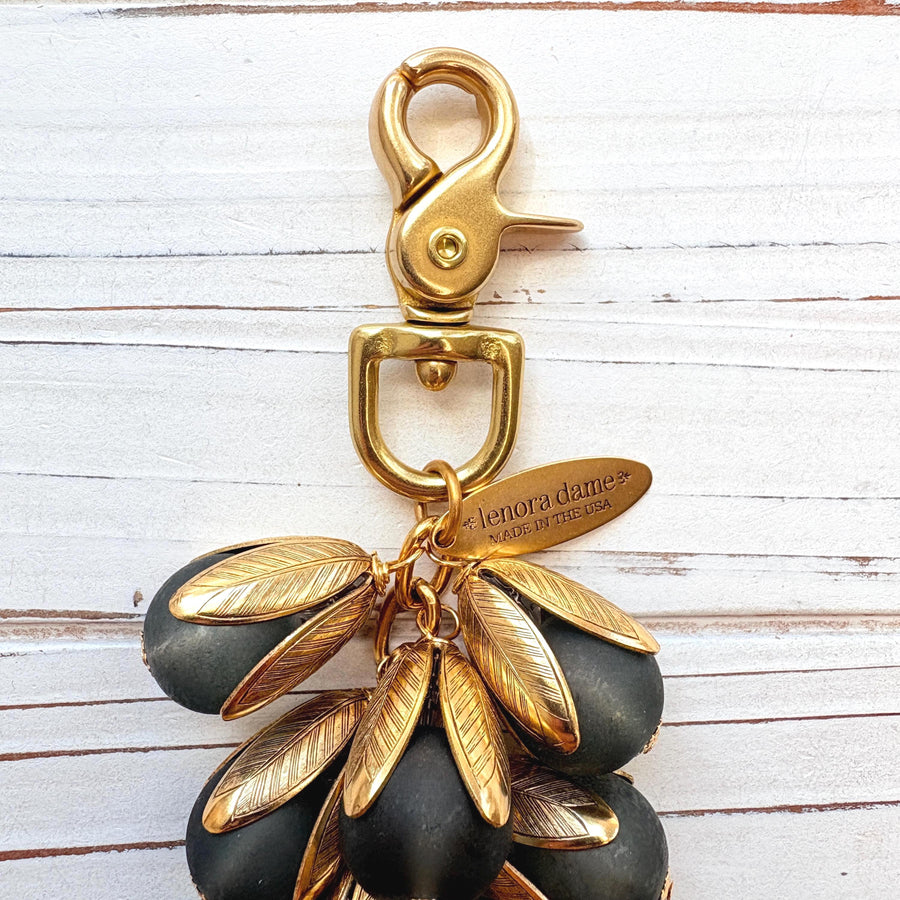 Beaded bag charm featuring matte black glass beads encased in gold plated brass floral leaf shaped bead caps. Gold trigger clasp and an oval hang tag that says Lenora Dame Made in USA. Lenora Dame bag charm.