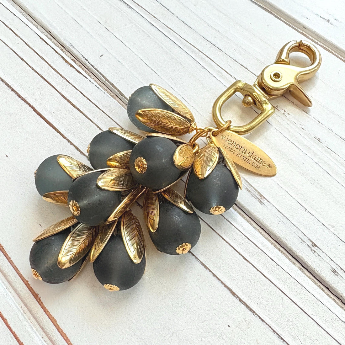 Beaded bag charm featuring matte black glass beads encased in gold plated brass floral leaf shaped bead caps. Gold trigger clasp and an oval hang tag that says Lenora Dame Made in USA. Lenora Dame bag charm.
