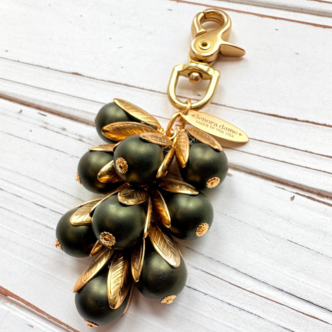 Gold bag charm with a trigger clasp at the top holding a cluster of matte hunter green pearls encased in gold leaf bead caps. A gold oval tag imprinted with Lenora Dame Handmade in USA hangs at the top by the trigger clasp. Lenora Dame bag charms. Green beaded purse charm.