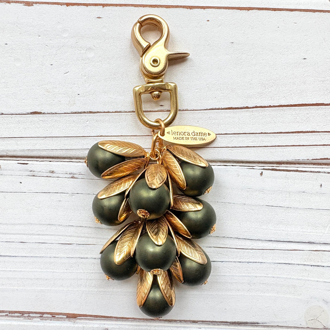 Gold bag charm with a trigger clasp at the top holding a cluster of matte hunter green pearls encased in gold leaf bead caps. A gold oval tag imprinted with Lenora Dame Handmade in USA hangs at the top by the trigger clasp. Lenora Dame bag charms. Green beaded purse charm.