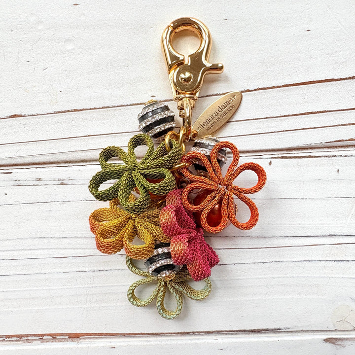 Floral bag charm with four mesh metal flowers in fall colors of green, orange and yellow, black and white striped beads with rhinestones and pumpkin orange pearls. Gold trigger clasp at the top with a gold oval charm that says Lenora Dame Made in USA.