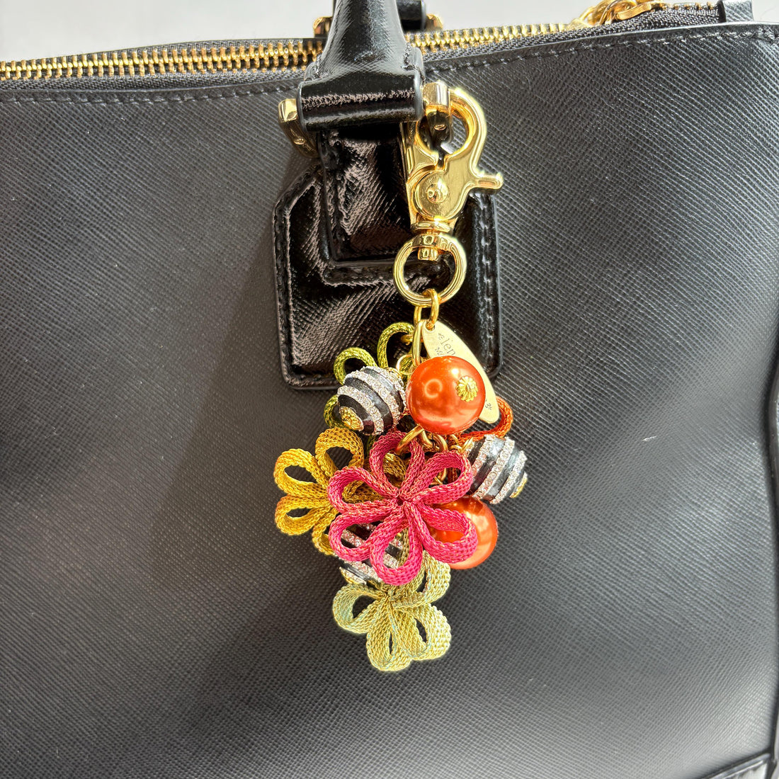 Floral bag charm with four mesh metal flowers in fall colors of green, orange and yellow, black and white striped beads with rhinestones and pumpkin orange pearls. Gold trigger clasp at the top with a gold oval charm that says Lenora Dame Made in USA.  Lenora Dame bag charms shown on the strap of a black leather handbag.