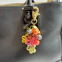 Floral bag charm with four mesh metal flowers in fall colors of green, orange and yellow, black and white striped beads with rhinestones and pumpkin orange pearls. Gold trigger clasp at the top with a gold oval charm that says Lenora Dame Made in USA.  Lenora Dame bag charms shown on the strap of a black leather handbag.