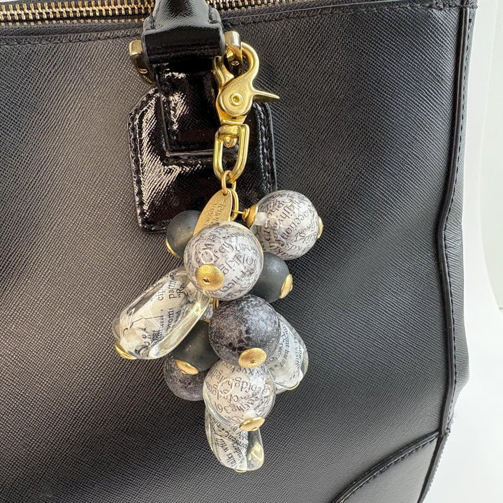 Irregular shaped beads with a newspaper print inside hang with round black foil beads and black glass beads in a cluster under a gold trigger clasp. Chunky bag charm is show hung on the strap of a black leather handbag. Lenora Dame bag charms.